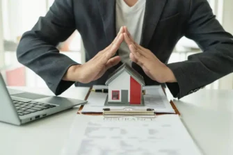 Man with hands in the shape of a roof over a model of a house for post about reviewing insurance.