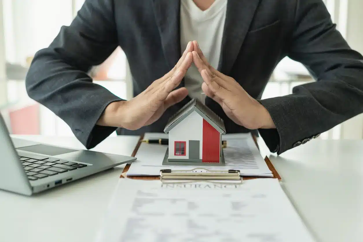 Man with hands folded in the shape of a roof over a model of a house to signify protecting a home with a periodic homeowners insurance review.